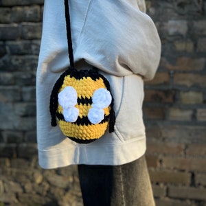 May include: A crocheted bee-shaped bag with black and yellow stripes and white wings. The bag is worn as a crossbody bag.