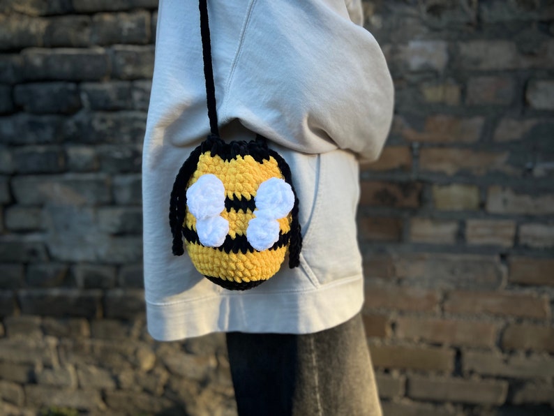 May include: A crocheted bee-shaped bag with black and yellow stripes and white wings. The bag is worn as a crossbody bag.