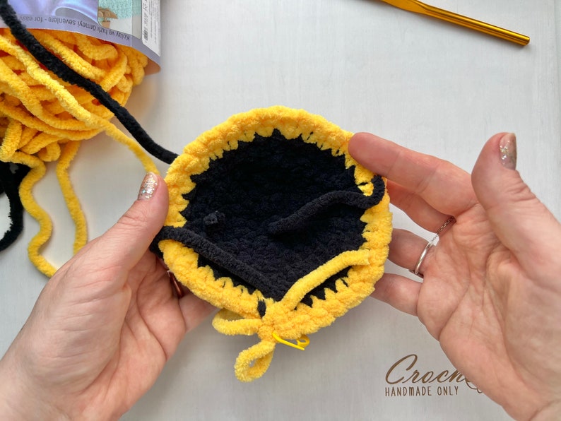 May include: A close-up of a crocheted yellow and black circle with a black center. The circle is being held by two hands, and the yellow yarn is being pulled through the center.