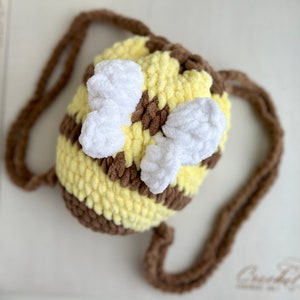May include: A crocheted bee-shaped bag with a brown strap. The bag is yellow and brown with white crocheted wings.