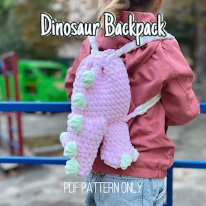 Op de afbeelding: Een roze, handgemaakte dinosaurusrugzak met mintgroene details. De rugzak heeft een afgeronde vorm en een zacht, getextureerd uiterlijk. De tekst "Dinosaur Backpack" staat bovenaan en "PDF PATTERN ONLY" onderaan.