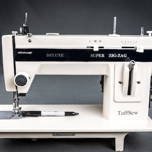 May include: A white TuffSew Deluxe Super Zig-Zag sewing machine with a black trim and a silver foot pedal. The machine has a needle, thread, and a bobbin.