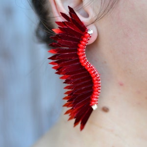 Op de afbeelding: Een paar rode en zilveren oorbellen in de vorm van een vleugel. De oorbellen zijn gemaakt van pailletten en kralen.