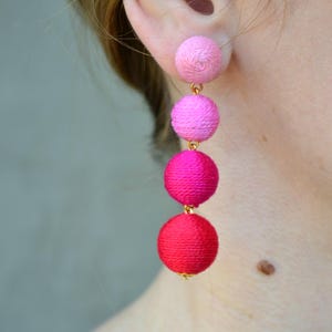 May include: Dangling earrings with three spheres in shades of pink and red. The top sphere is light pink, followed by medium pink, and a deep red sphere at the bottom. Gold-coloured connectors link the spheres.