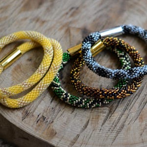 Peut inclure: Un ensemble de quatre bracelets circulaires en perles aux motifs complexes. Un bracelet est jaune avec des fermoirs en métal doré, tandis que les autres présentent des perles vertes, brunes et noires avec des fermoirs cylindriques argentés et dorés.