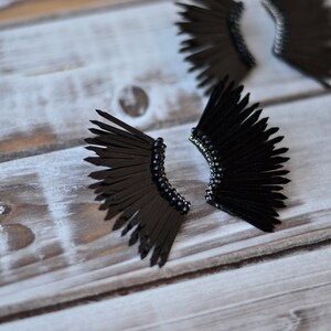 May include: A pair of black feather earrings with a beaded detail. The earrings are shaped like wings and are made of black feathers.
