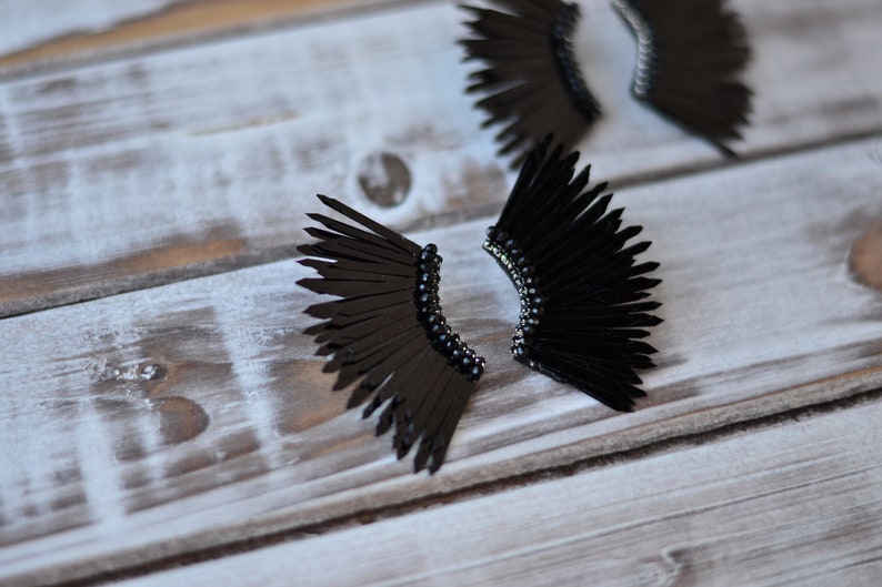 May include: A pair of black feather earrings with a beaded detail. The earrings are shaped like wings and are made of black feathers.