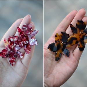 May include: A pair of statement earrings featuring a floral design. The earrings are made of a tortoiseshell-like material and are available in two colour variations: red and white, and brown and black.