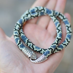 以下が含まれることがあります： 銀色の蛇の頭の留め具が付いたビーズの蛇のネックレス。ネックレスは黒、白、青のビーズで作られています。