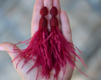May include: A pair of long, deep red earrings featuring a beaded stud top connected to a smaller beaded section, which transitions into a long, flowing fringe of soft, delicate feathers in a matching dark red color.
