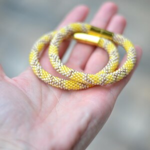 以下が含まれることがあります： 鮮やかな黄色とメタリックシルバーの斜めストライプが交互に並ぶ、小さなビーズを密に編み込んだ円形のブレスレット。両端を固定する磨き上げられたゴールドトーンの円筒形マグネットクラスプが特徴です。