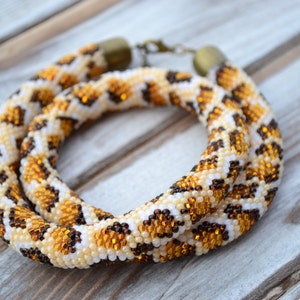 May include: A beaded necklace with a brown, white, and gold leopard print pattern. The necklace is made of small beads and is coiled on a wooden surface.