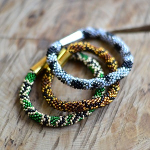 May include: Three beaded bracelets in different patterns. The first bracelet is black and white with silver beads. The second bracelet is brown and gold with black beads. The third bracelet is green, gold, and black.