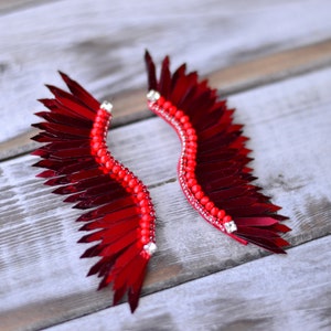 Op de afbeelding: Een paar rode veren oorbellen met een pareldetail. De veren zijn in de vorm van een vleugel gerangschikt en zijn versierd met kleine, heldere strass steentjes.