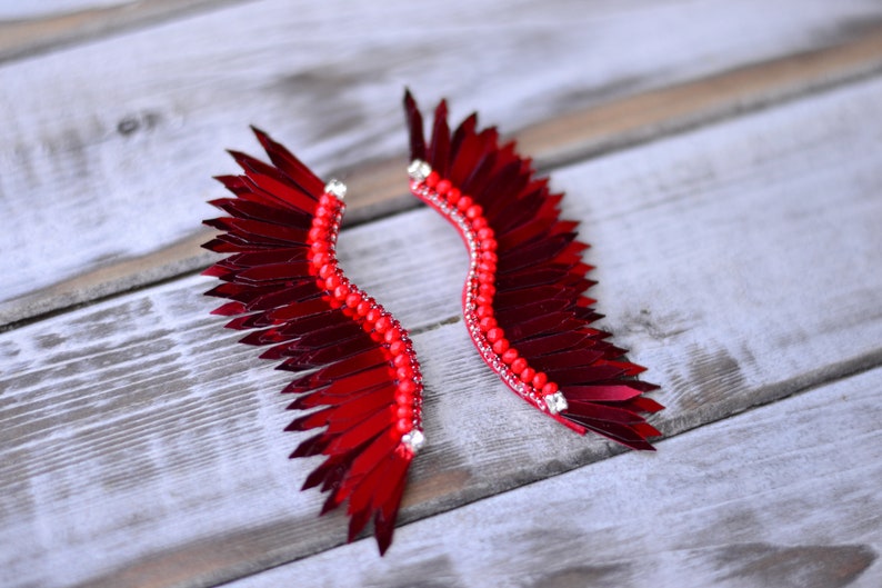 Op de afbeelding: Een paar rode veren oorbellen met een pareldetail. De veren zijn in de vorm van een vleugel gerangschikt en zijn versierd met kleine, heldere strass steentjes.