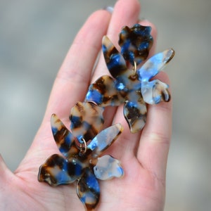 以下が含まれることがあります： 青と茶色の花柄のデザインが施された、大きなステートメントイヤリングのペア。イヤリングは、大理石模様の透き通った素材で作られています。