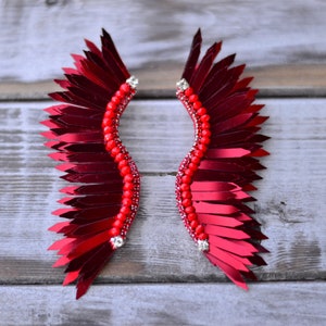 Op de afbeelding: Een paar rode veren oorbellen met een parelontwerp. De oorbellen zijn gevormd als vleugels en hebben een glanzende, metallic afwerking.