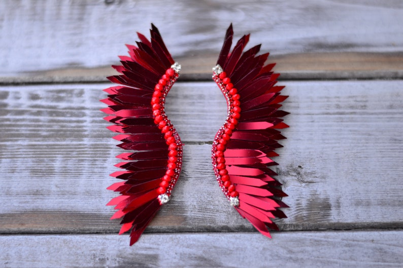 Op de afbeelding: Een paar rode veren oorbellen met een parelontwerp. De oorbellen zijn gevormd als vleugels en hebben een glanzende, metallic afwerking.