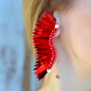 Op de afbeelding: Een grote, rode, veerachtige oorbel met een zilveren accent. De oorbel is gemaakt van veel kleine, overlappende stukjes stof.