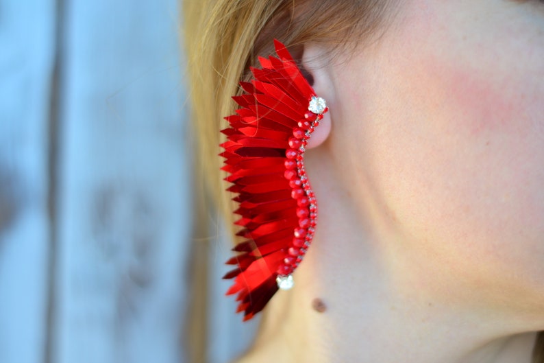 Op de afbeelding: Een grote, rode, veerachtige oorbel met een zilveren accent. De oorbel is gemaakt van veel kleine, overlappende stukjes stof.