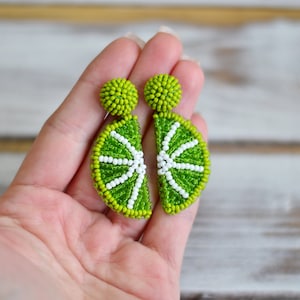 May include: A pair of lime green and white beaded earrings shaped like lime slices. The earrings are held in a hand.