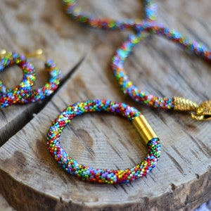 May include: A set of colourful beaded jewellery, including a necklace, bracelet, and earrings. The jewellery is made with small, multi-coloured beads and features gold accents.