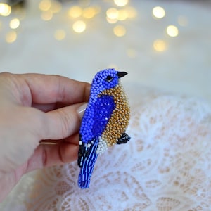 May include: A beaded bluebird brooch. The bird features a blue head and wings, a golden-brown body, and black and white accents. The brooch is made of small beads and is held in a hand.