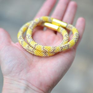 以下が含まれることがあります： 鮮やかな黄色とメタリックシルバーのシードビーズが交互に並ぶ、柔軟なチューブ状のビーズブレスレット。光沢のあるゴールドトーンの円筒形マグネットクラスプが2つ付いており、それらが連結して途切れのない輪を形成しています。