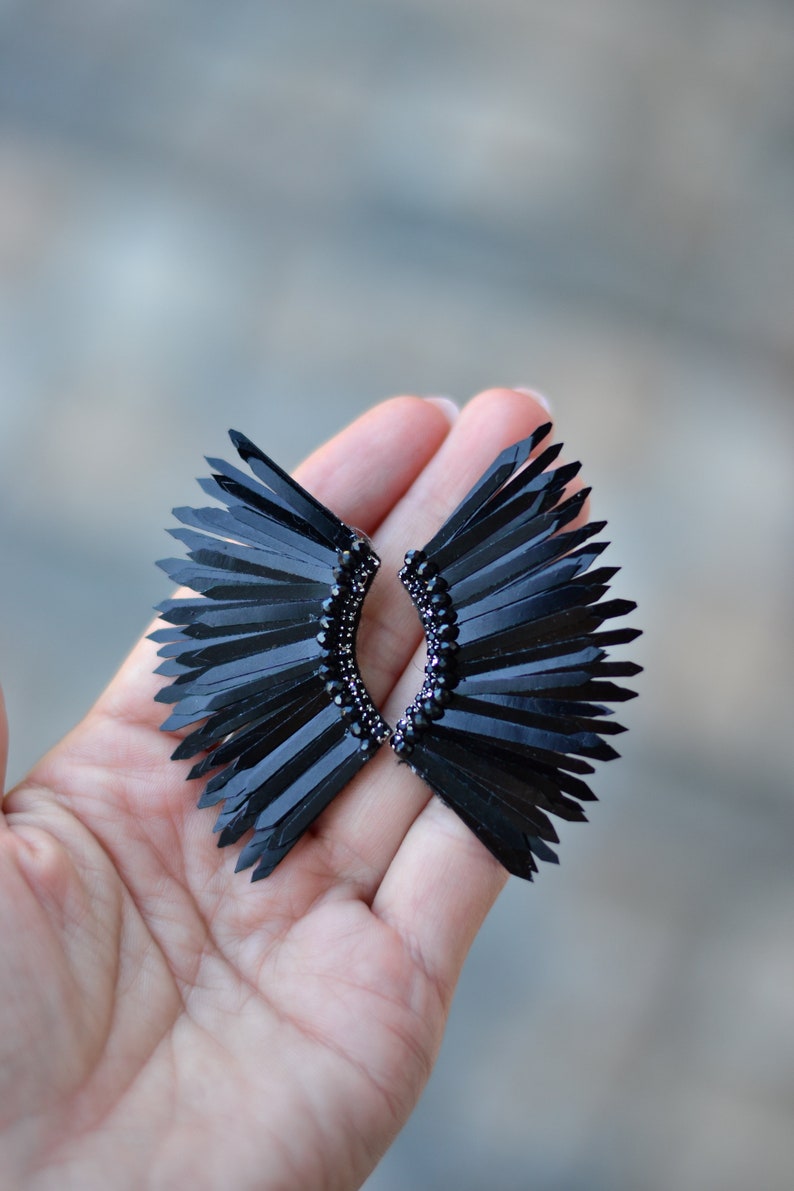 May include: A pair of black earrings with a fan-shaped design. The earrings are made of black feathers and have a black bead detail.