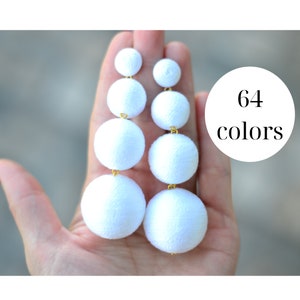 May include: White statement earrings with four round pom-poms, each decreasing in size. The earrings are made of fabric and have a gold-tone metal connector. 64 colors.
