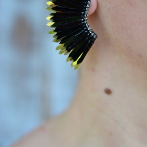 May include: A pair of black and gold earrings with a fan-like design. The earrings are made of metal and have a geometric shape.