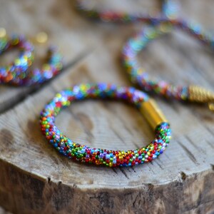 May include: Three colourful beaded bracelets with gold accents. The bracelets are made of small, colourful beads and are arranged on a wooden surface.
