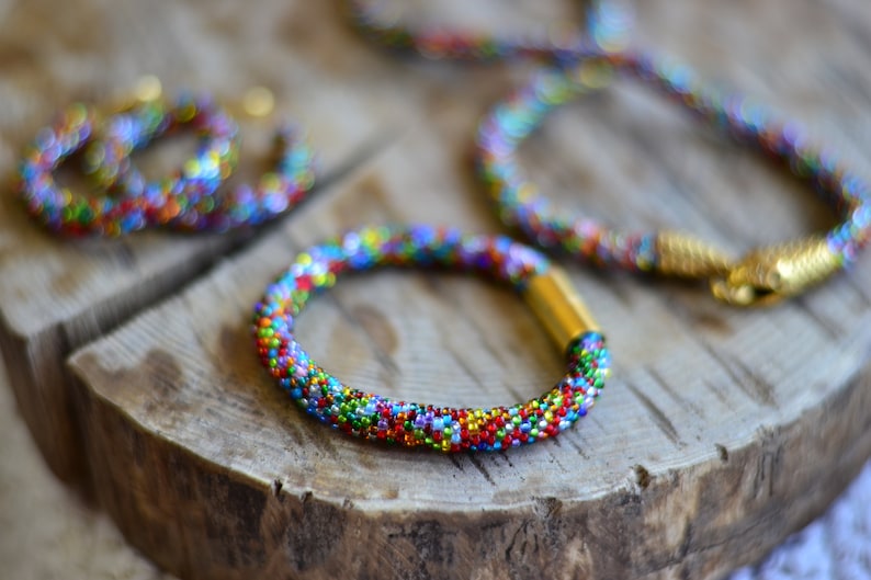 May include: Three colourful beaded bracelets with gold accents. The bracelets are made of small, colourful beads and are arranged on a wooden surface.
