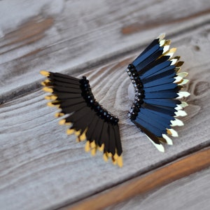 May include: A pair of black and gold earrings with a fan-like design. The earrings are made of black feathers with gold tips and have black beads around the base.