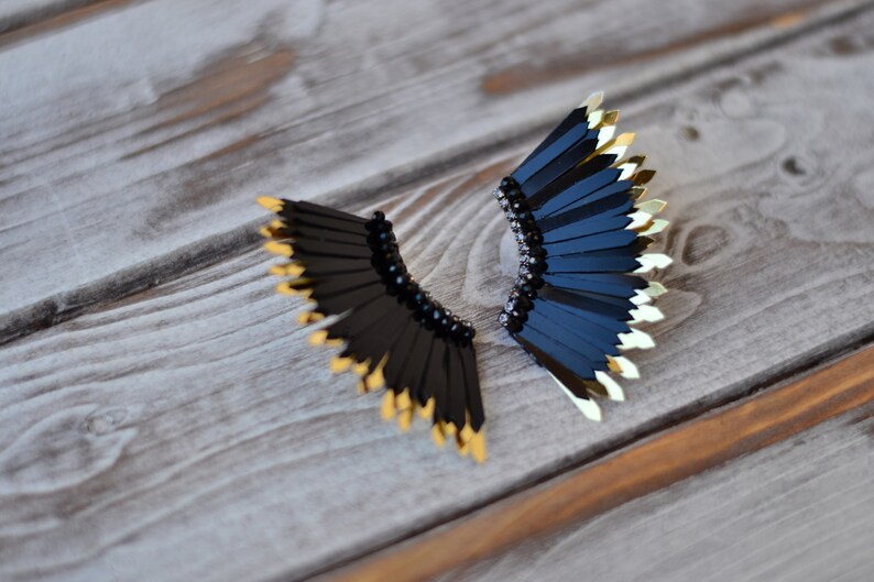 May include: A pair of black and gold earrings with a fan-like design. The earrings are made of black feathers with gold tips and have black beads around the base.