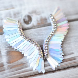 May include: A pair of iridescent angel wings made of sequins and rhinestones. The wings are a light blue and white colour and are arranged in a symmetrical pattern.