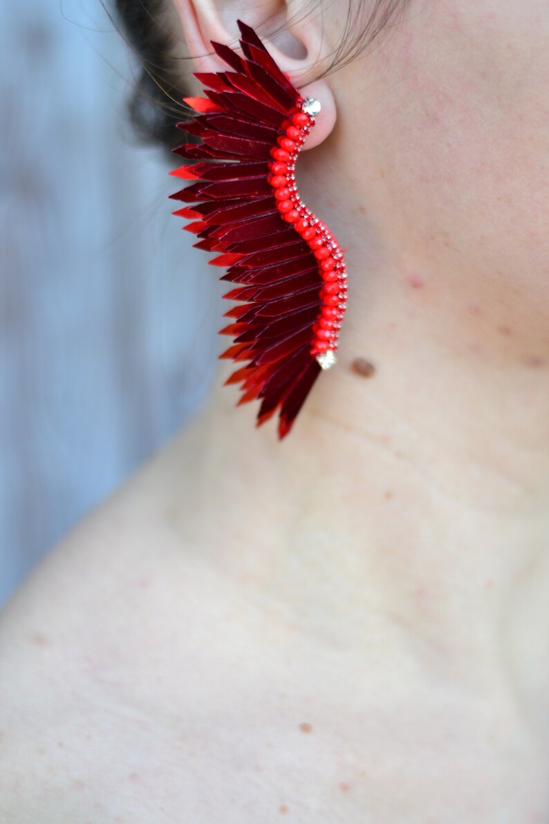 Op de afbeelding: Een paar rode en zilveren oorbellen in de vorm van een vleugel. De oorbellen zijn gemaakt van pailletten en kralen.