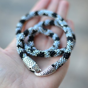 May include: A black and silver beaded necklace with a snake head clasp. The necklace is coiled and displayed on a hand.
