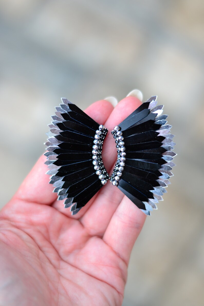 May include: A pair of black and silver earrings with a fan-like design. The earrings are made of small, black and silver beads that are arranged in a fan shape. The earrings are held together by a silver wire.