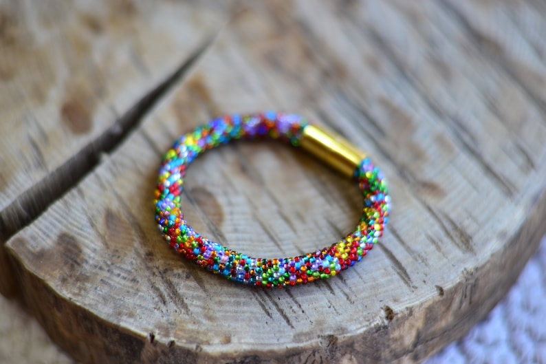 May include: A colourful beaded bracelet with a gold clasp. The bracelet is made of small, multi-coloured beads and is woven in a tight pattern.