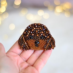 May include: A dog-shaped brooch made of brown and bronze beads. The brooch has black beads for the eyes and nose. The dog's fur is detailed with various shades of brown beads, creating a textured appearance.