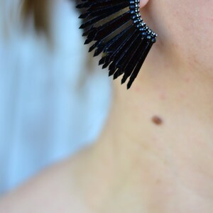 May include: A pair of black statement earrings with a fan-shaped design. The earrings are made of multiple thin, black pieces that are arranged in a fan shape. The earrings are large and dramatic.