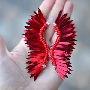 Op de afbeelding: Een paar rode veren oorbellen met een parelmoer centrum. De oorbellen zijn gevormd als vleugels en hebben een glanzende, metallic afwerking.