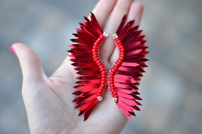 Op de afbeelding: Een paar rode veren oorbellen met een parelmoer centrum. De oorbellen zijn gevormd als vleugels en hebben een glanzende, metallic afwerking.