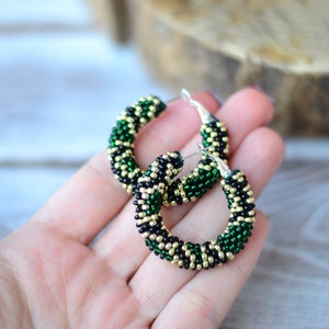 May include: A pair of green, black and gold beaded hoop earrings. The earrings are made with small beads and have a silver hook fastening.