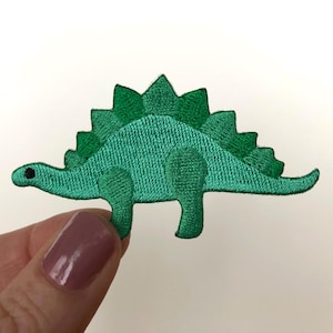 May include: Green embroidered iron-on patch of a cartoon dinosaur with a spiky back.