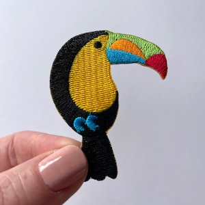 May include: Embroidered iron-on patch of a toucan with a yellow body, black head and tail, and a multi-coloured beak. The beak is green, orange, blue, and red.
