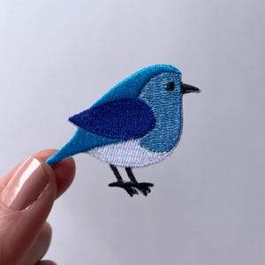 May include: An embroidered bird patch in blue and white. The bird has a white belly and a blue body with a darker blue wing.