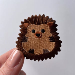 May include: A brown embroidered iron-on patch of a cartoon hedgehog with a brown nose and black eyes.