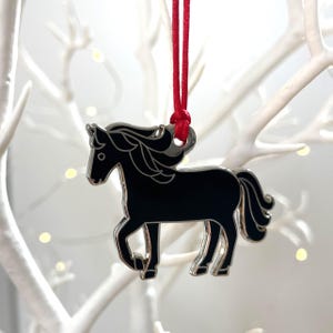 May include: A black horse ornament with silver edging, suspended by a red cord. The horse is depicted in a trotting stance, with a flowing mane and tail. The ornament is likely metal and is a decorative item.